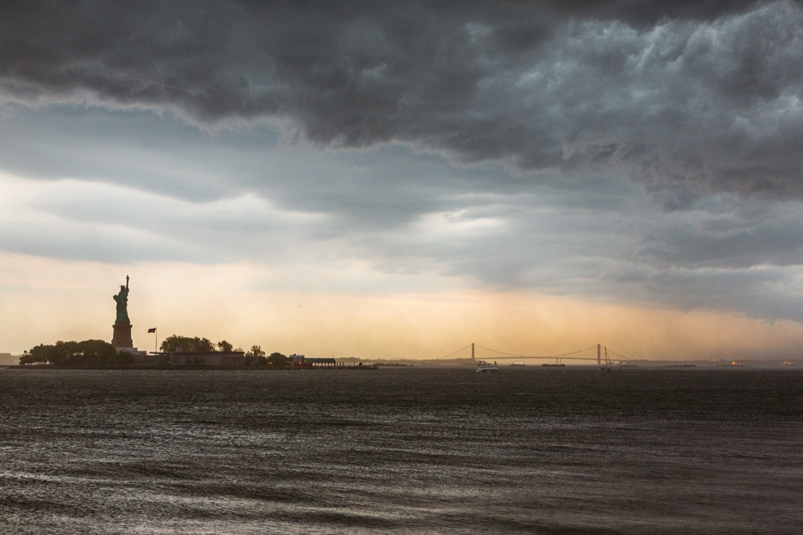 The Statue of Liberty on the horizon under heavy storm clouds, with a band of golden light over the water.