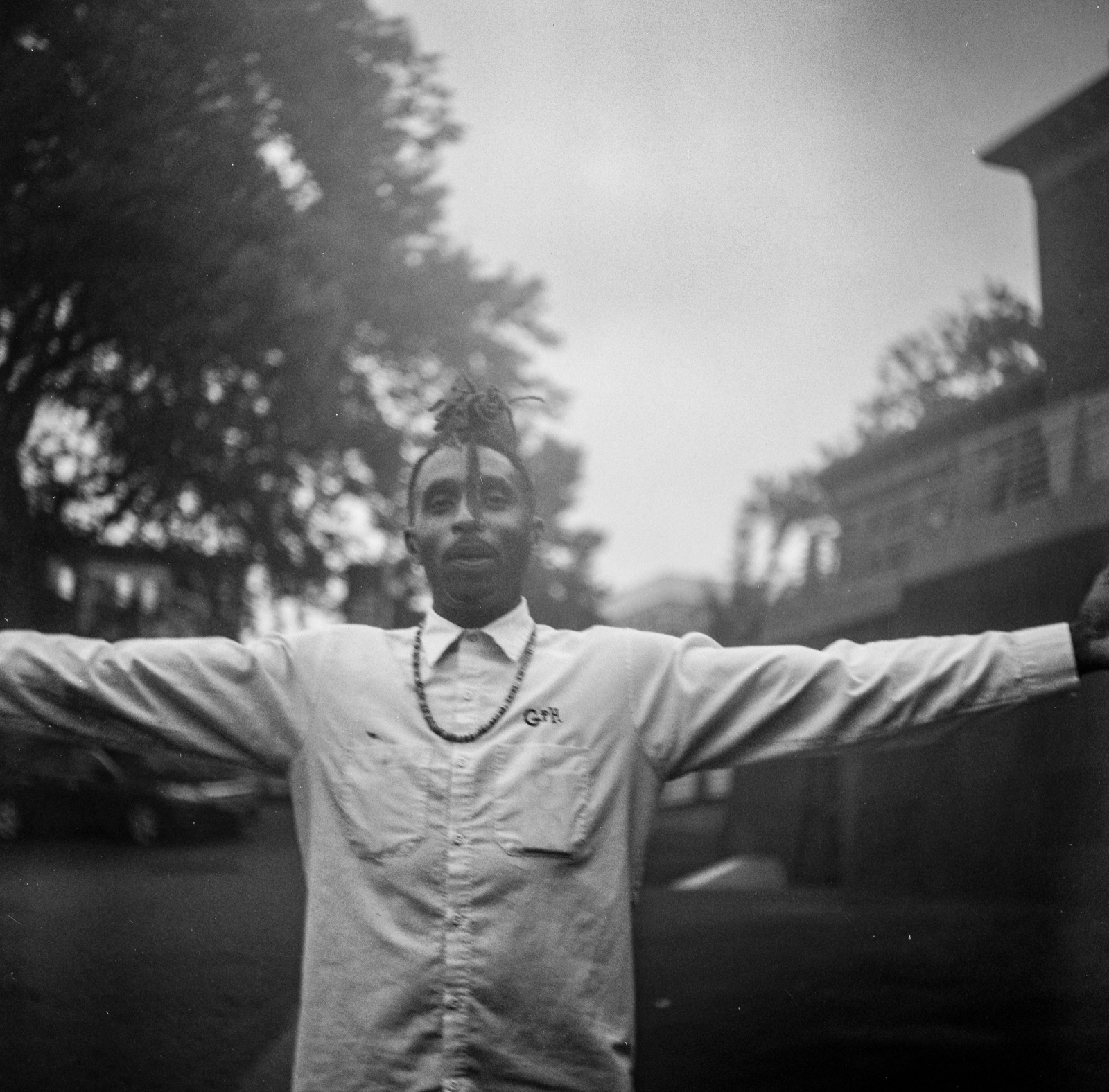 Black-and-white portrait of a man in a work shirt and bead necklace, arms stretched wide on a leafy street.